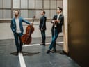Drei Männer in Anzügen mit Musikinstrumenten in einem modernen Raum.