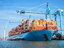 Grosses Containerschiff im Hafen mit blauen Kränen und Windkraftanlage im Hintergrund.