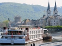 Flusskreuzfahrtschiff vor einer historischen Stadt mit Türmen und Hügeln im Hintergrund.