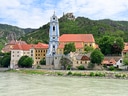 Blick auf die Stadt Dürnstein mit Barockkirche und Burgruine, umgeben von Hügeln an der Donau.