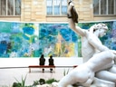 Zwei Personen betrachten ein grosses farbenfrohes Gemälde neben einer weissen Statue im Museum.
