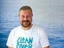 Mann im T-Shirt mit 'Clean River'-Aufdruck vor Wasserhintergrund.