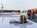 Weinglas und Fernglas auf einem Tisch im Freien, im Hintergrund Menschen in Stühlen.