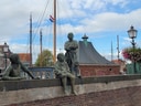 Bronzeskulpturen von drei Kindern auf einer Backsteinmauer mit Segelmasten im Hintergrund.