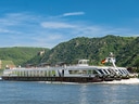 Flusskreuzfahrtschiff auf dem Rhein vor grüner Hügelkulisse.