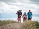 Drei Personen wandern auf einem sandigen Weg unter bewölktem Himmel.