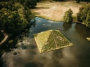 Luftaufnahme einer grasbewachsenen Pyramideninsel in einem See mit umliegendem Wald.