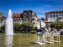 Springbrunnen mit Pferdestatuen vor Gebäuden.