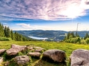 Aussicht über einen See von einer Wiese mit Felsen, im Hintergrund bewaldete Hügel und blauer Himmel.