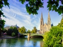 Kirchengebäude mit zwei Türmen hinter Fluss und Brücke, umgeben von Bäumen.