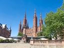 Neugotische Kirche mit roten Türmen und Statue im Vordergrund.