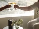 Zwei Personen stossen mit Weingläsern an einem Tisch an, im Hintergrund Wasserblick.
