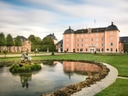 Elegantes Schloss mit rosa Fassade und Kuppeldächern, umgeben von gepflegten Gärten und einem Teich mit Statue im Vordergrund.