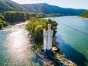 Alte Burg am Flussufer mit bewaldeten Hügeln im Hintergrund.