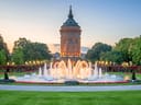 Wasserturm mit beleuchtetem Brunnen und Gärten bei Sonnenuntergang.