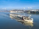 Grosses Kreuzfahrtschiff fährt auf einem Fluss mit bewaldeten Hügeln im Hintergrund.