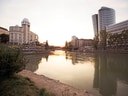 Flussufer bei Sonnenuntergang mit modernen Gebäuden im Hintergrund.