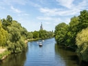 Fluss mit Booten und Kirchturm im Hintergrund, umgeben von Bäumen.
