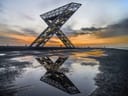 Grosse Stahlskulptur bei Sonnenuntergang mit Pfützen im Vordergrund.