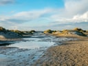 Strandlandschaft mit Dünen und Wasserpfützen bei bewölktem Himmel.