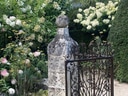 Steingartenstatue mit kunstvollem Tor und blühenden Sträuchern im Hintergrund.