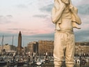 Statue eines Mannes mit einem Kind auf den Schultern vor einem Hafen mit Booten.