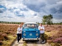 Zwei Personen lehnen an einem blauen Retro-Van auf einem ländlichen Feldweg mit violetten Blumen.