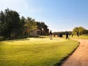 Mehrere Personen stehen auf einem Golfplatz in der Nähe eines Holzschuppens bei sonnigem Wetter.