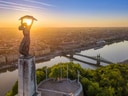 Freiheitsstatue auf Hügel mit Fluss und Stadt im Sonnenuntergang.