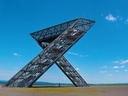 Grosse Metallskulptur in Form eines 'X' vor klarem Himmel.