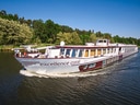 Kreuzfahrtschiff fährt auf Fluss vor bewaldeter Kulisse.