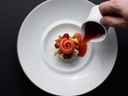 Nahaufnahme eines kunstvoll angerichteten Desserts auf einem weissen Teller, darüber rote Sauce aus einer weissen Kanne.
