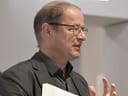 Person in schwarzem Anzug hält Papiere und gestikuliert während des Sprechens.