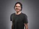 Mann mit Brille und lockigem Haar lächelt vor grauem Hintergrund.