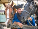 Person in traditioneller Kleidung mit zwei Pferden in einem Wagen.