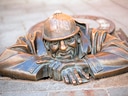 Bronzestatue eines Mannes, der aus einem Gully schaut.