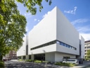 Weisse moderne Gebäude an einer Strassenecke mit Bäumen und vorbeifahrenden Autos.