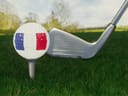 Ein Golfball mit Flaggenmuster auf einem Tee vor einem Golfschläger auf dem Rasen.