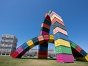 Grosse Skulptur aus bunten Containern auf einer Wiese vor einem klaren Himmel.
