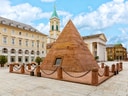 Platz mit roter Pyramide, von Gebäuden umgeben, darunter eine Kirche mit Turm.