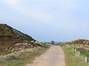 Weg zwischen sandigen Dünen mit Radfahrern in der Ferne.