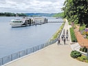 Radfahrer auf einem Uferweg neben einem Fluss mit einem angelegten Schiff.