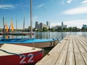 Hölzerner Steg am Wasser mit kleinen Segelbooten und Stadtansicht im Hintergrund.