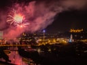 Nächtliche Stadtlandschaft mit Feuerwerk am Himmel, beleuchteten Gebäuden und Fluss.