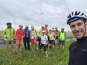 Gruppe von Radfahrern in bunter Kleidung posiert auf einem Feldweg mit Windrädern im Hintergrund.