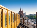 Gelber Zug auf Hochbahnstrecke mit historischen Türmen im Hintergrund.