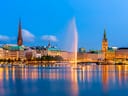 Stadtansicht mit Gebäuden und Springbrunnen am Wasser bei Abenddämmerung.