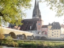 Drei Personen sitzen am Fluss vor der Steinernen Brücke und dem Dom in Regensburg.