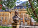 Bronzebüste vor einem historischen Gebäude mit Bäumen und Fahnen.