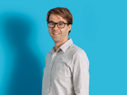 Person mit Brille und hellem Hemd vor blauem Hintergrund.